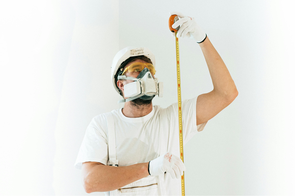 Construction worker wearing protective gear measuring a wall with a tape measure.