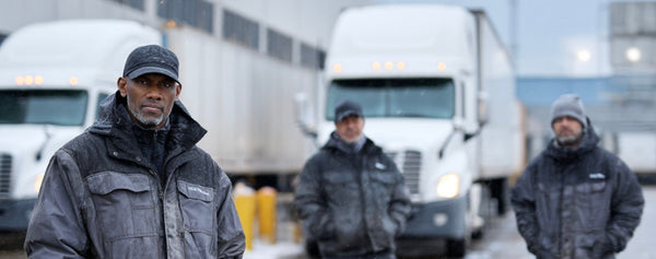 Industrial workers standing outdoors in winter conditions with delivery trucks in the background.